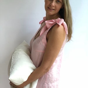May include: A light pink linen sleep dress with bow details on the shoulders. The dress has a V-neck and is sleeveless. A white pillow is held in the arms. The dress is made of a soft, breathable fabric.