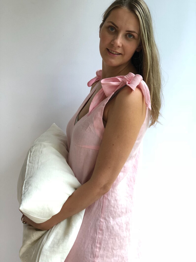 May include: A light pink linen sleep dress with bow details on the shoulders. The dress has a V-neck and is sleeveless. A white pillow is held in the arms. The dress is made of a soft, breathable fabric.