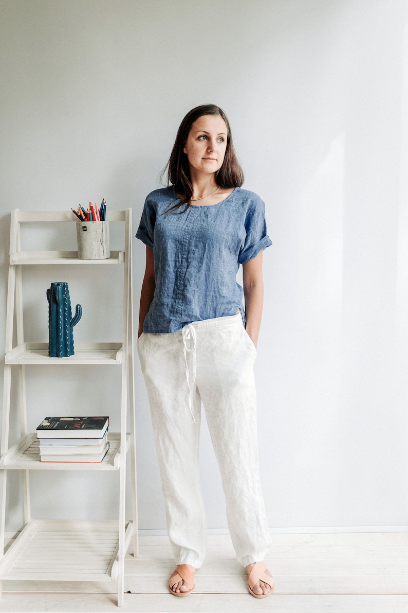 May include: A woman wearing a blue linen shirt and white linen pants. She is standing in front of a white shelf with a cactus and a pencil holder on it.