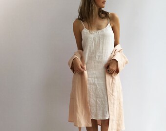 Op de afbeelding: Een vrouw draagt een witte linnen slipdress met een roze linnen badjas over haar schouders. Ze staat op een lichte houten vloer.