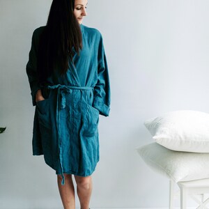 May include: A woman wearing a teal blue linen robe with a sash tie and pockets. She is standing on a white wooden floor and wearing brown sandals.
