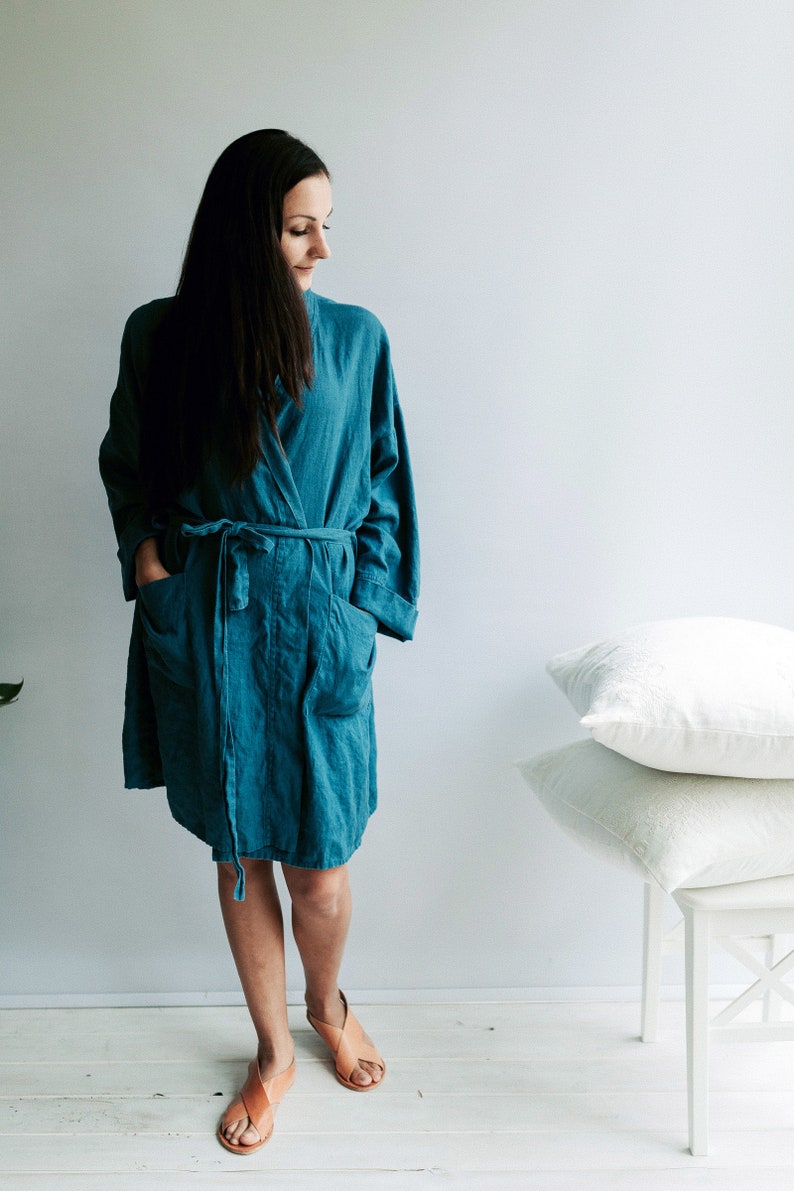 May include: A woman wearing a teal blue linen robe with a sash tie and pockets. She is standing on a white wooden floor and wearing brown sandals.
