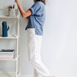 May include: A woman wearing a light blue linen shirt and white linen pants. She is standing next to a white shelf with a cactus and books on it. She is holding a red pencil in her right hand.