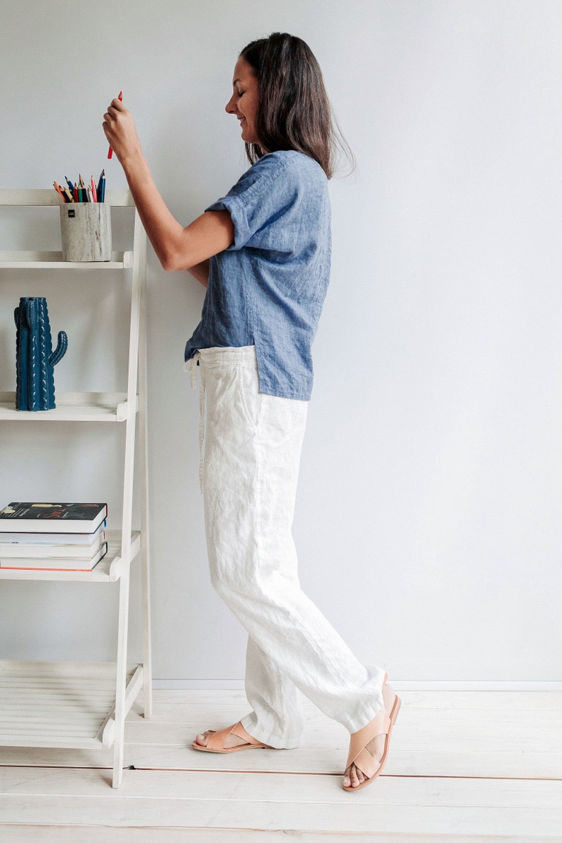 May include: A woman wearing a light blue linen shirt and white linen pants. She is standing next to a white shelf with a cactus and books on it. She is holding a red pencil in her right hand.