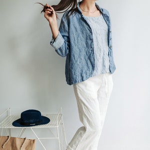 May include: A woman wearing a light blue linen shirt jacket over a white and blue striped shirt, white linen pants, and peach-colored sandals. She is holding her long brown hair in a ponytail. A blue hat and a brown paper bag are on a white metal shelf in the background.