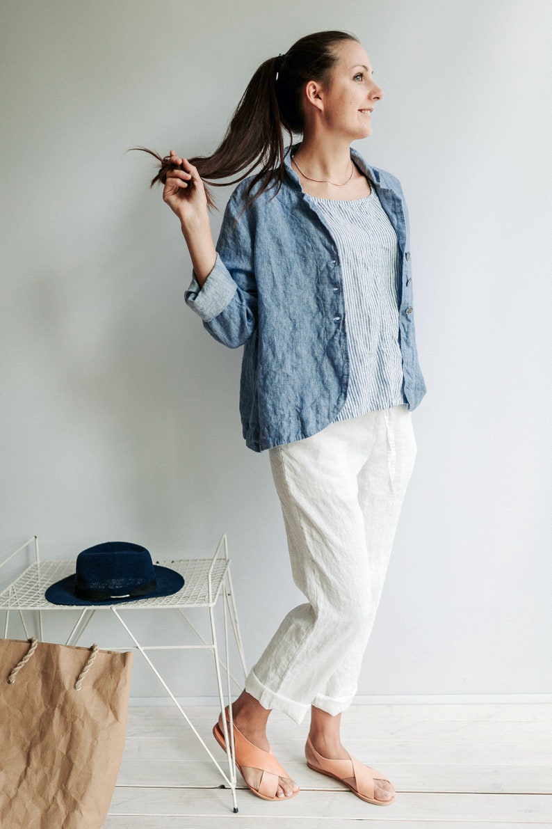 May include: A woman wearing a light blue linen shirt jacket over a white and blue striped shirt, white linen pants, and peach-colored sandals. She is holding her long brown hair in a ponytail. A blue hat and a brown paper bag are on a white metal shelf in the background.