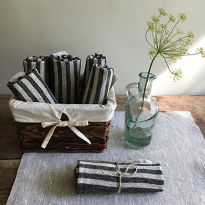 May include: A wicker basket with a white fabric lining holds several rolled black and white striped towels. A small bundle of similar towels is tied with twine. A clear glass bottle with a single flower is also present.