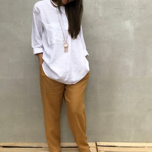 K&ouml;nnte beinhalten: Eine Frau tr&auml;gt ein wei&szlig;es Leinenhemd mit einer goldenen Halskette und einem Strohhut. Sie tr&auml;gt eine senfgelbe Leinenhose und braune Sandalen. Sie steht auf einer Holzpalette.