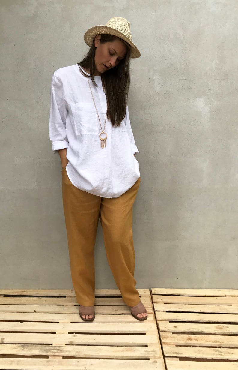 K&ouml;nnte beinhalten: Eine Frau tr&auml;gt ein wei&szlig;es Leinenhemd mit einer goldenen Halskette und einem Strohhut. Sie tr&auml;gt eine senfgelbe Leinenhose und braune Sandalen. Sie steht auf einer Holzpalette.