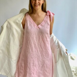 May include: A light pink linen dress with a V-neck and bow details on the shoulders. The dress is sleeveless and falls to the mid-thigh. A white robe is draped over the shoulders.