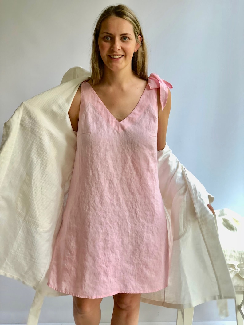 May include: A light pink linen dress with a V-neck and bow details on the shoulders. The dress is sleeveless and falls to the mid-thigh. A white robe is draped over the shoulders.