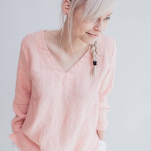 May include: A pale pink, long-sleeved linen blouse with a v-neckline. The blouse is made of a soft, lightweight fabric and has a relaxed fit.