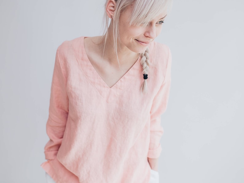 May include: A pale pink, long-sleeved linen blouse with a v-neckline. The blouse is made of a soft, lightweight fabric and has a relaxed fit.