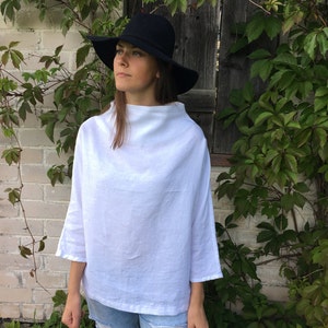Puede incluir: Una blusa blanca de lino con cuello amplio de barco y mangas 3/4. La blusa es holgada y tiene un estilo relajado y casual. La modelo lleva la blusa con jeans azules y un sombrero negro de ala ancha.
