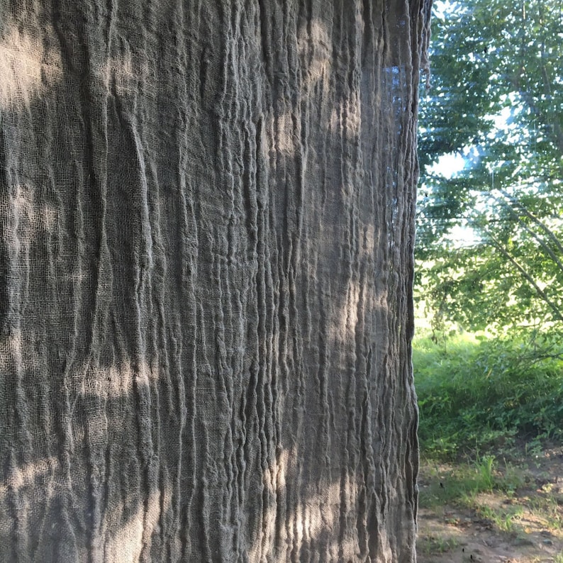 Wrinkled linen curtains hanging by a window. Light filters through the gauzy wrinkled fabric and shadows from the nearby trees are playing on the fabric.