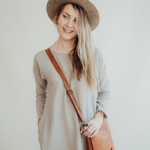 May include: A woman wearing a light gray linen dress with a brown leather crossbody bag and a wide-brimmed gray hat.