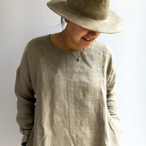 A woman is wearing a natural color linen tunic with long sleeves. Her hands are placed in pockets. The tunic features a comfortable cut.