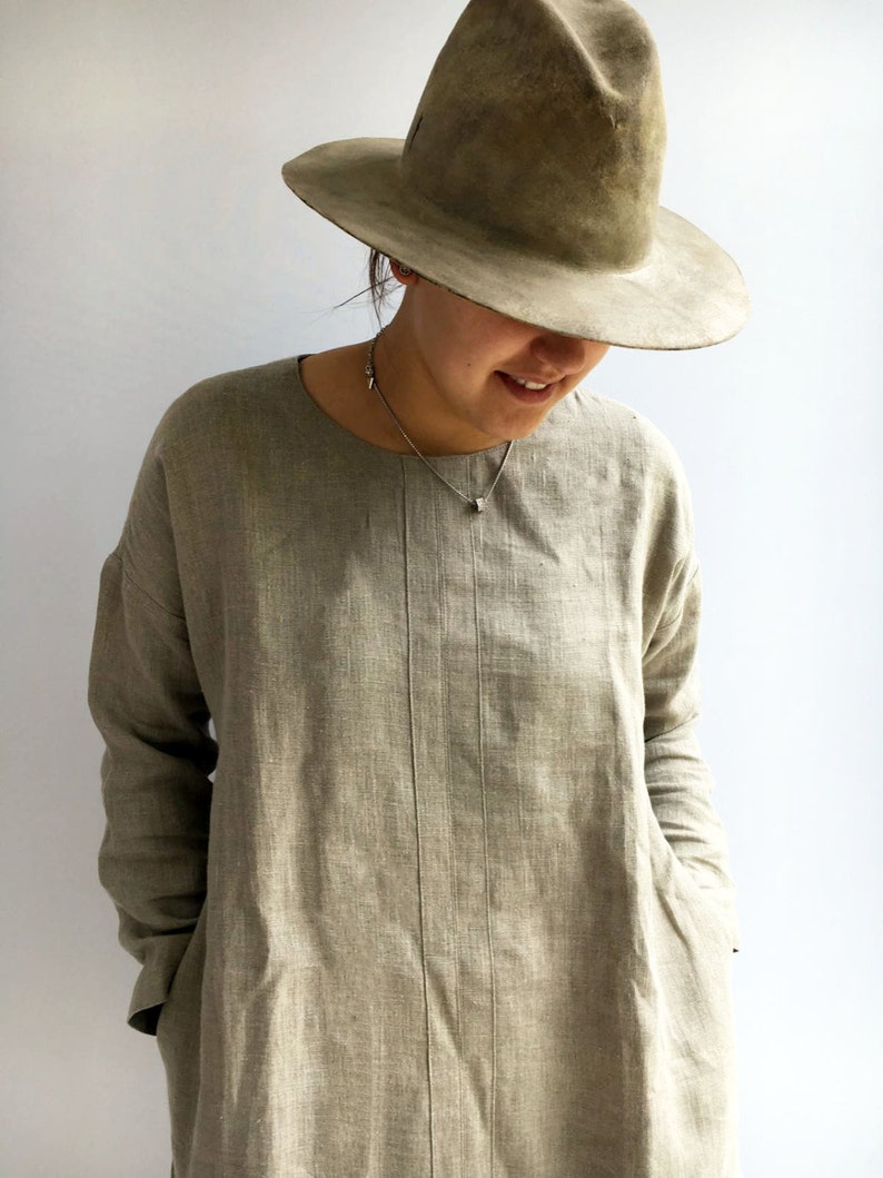 A woman is wearing a natural color linen tunic with long sleeves. Her hands are placed in pockets. The tunic features a comfortable cut.