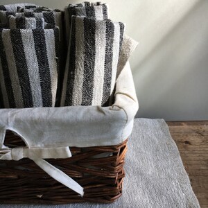 May include: A woven wicker basket filled with rolled, striped fabric. The fabric has alternating black and off-white vertical stripes. The basket is lined with a cream-colored fabric and sits on a gray linen surface.