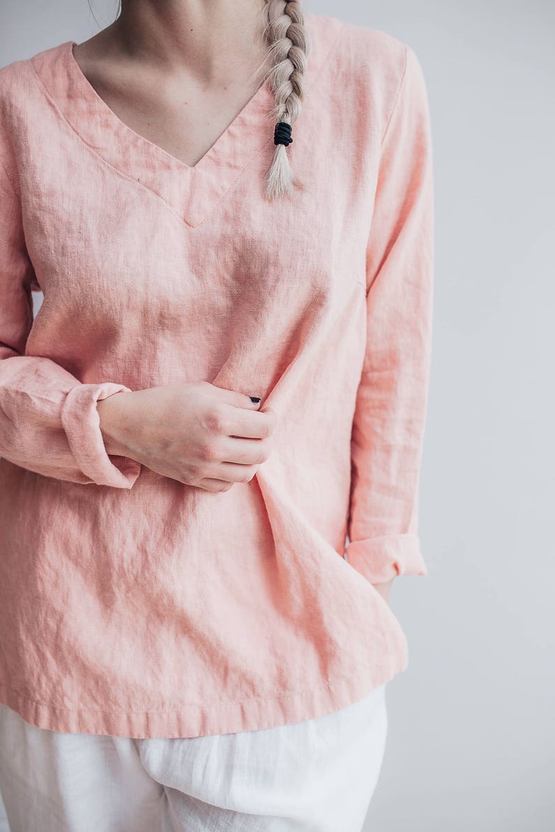 May include: A pale pink linen blouse with a v-neck and long sleeves. The blouse is loose fitting and has a relaxed, casual style.