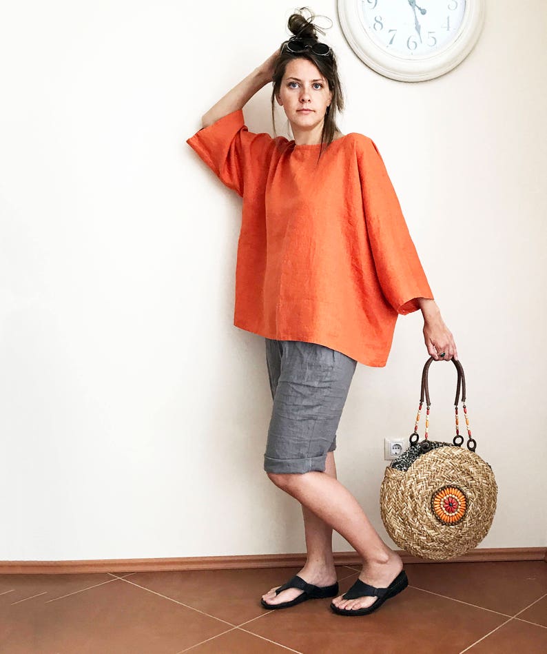 K&ouml;nnte beinhalten: Eine Frau tr&auml;gt ein locker sitzendes, orangefarbenes Leinenoberteil mit grauen Leinenhosen und schwarzen Sandalen. Sie h&auml;lt eine runde, geflochtene Strohtasche mit einem braunen Ledergriff und bunten Perlen.
