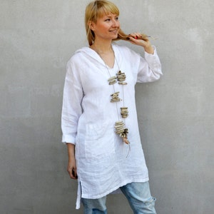 Op de afbeelding: Een vrouw draagt een witte linnen tuniek met een lange, gelaagde ketting gemaakt van drijfhout. Ze draagt een blauwe jeans met een distressed look en staat voor een grijze muur.