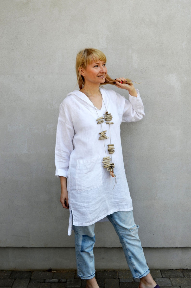 Op de afbeelding: Een vrouw draagt een witte linnen tuniek met een lange, gelaagde ketting gemaakt van drijfhout. Ze draagt een blauwe jeans met een distressed look en staat voor een grijze muur.