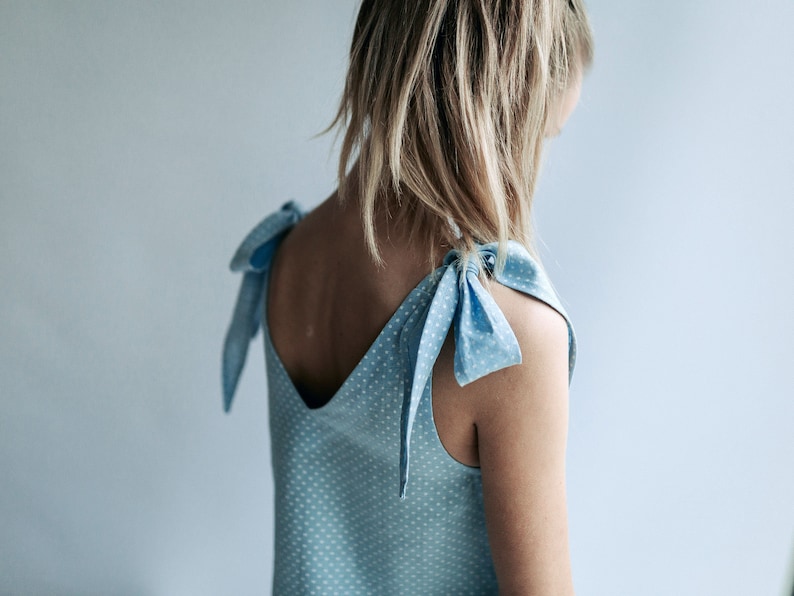 May include: Light blue sleeveless top with white polka dots and tie shoulder straps. The top has a V-neck and is made of a lightweight fabric. The straps are tied into bows. The overall style is casual and feminine.