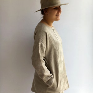 A woman is standing pictured in profile. She is wearing natural color comfortable linen tunic. The tunic has long sleeves and pockets. It runs over woman's hips. She is wearing jeans underneath.