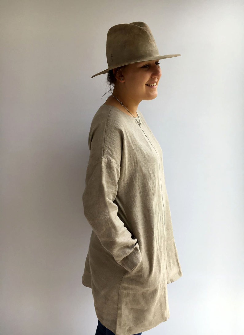 A woman is standing pictured in profile. She is wearing natural color comfortable linen tunic. The tunic has long sleeves and pockets. It runs over woman's hips. She is wearing jeans underneath.