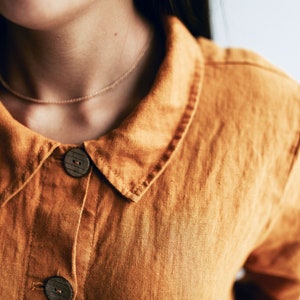 May include: A close-up of a brown linen shirt with a collar and two wooden buttons. The shirt has a simple, casual design.