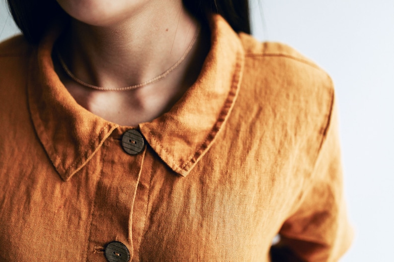 May include: A close-up of a brown linen shirt with a collar and two wooden buttons. The shirt has a simple, casual design.