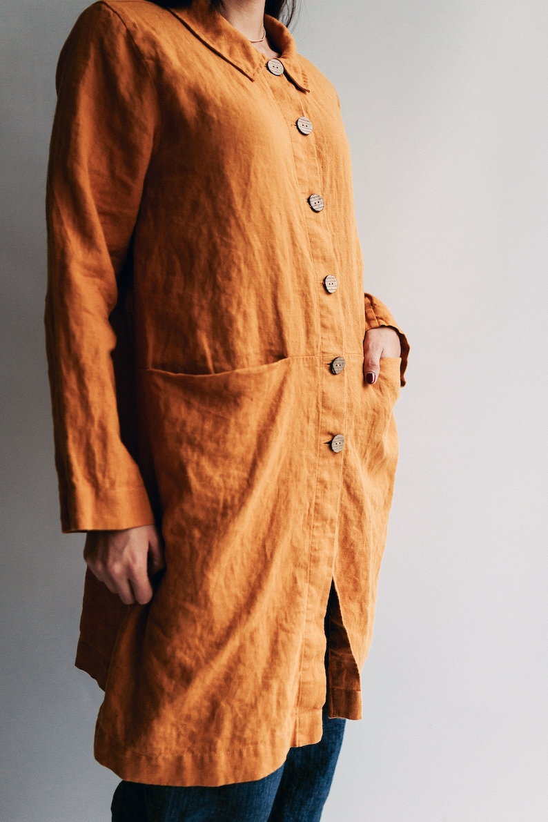 May include: A long, loose-fitting, mustard yellow linen shirt with a collar and button closure. The shirt has a single patch pocket on the left side and a slit in the back.