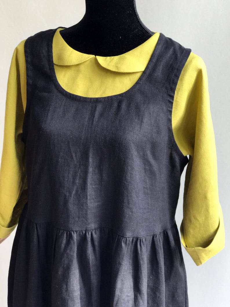 May include: A black linen dress with a gathered skirt and a Peter Pan collar. The dress is layered over a yellow collared shirt.