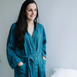 May include: A woman wearing a teal blue linen robe with a sash tie. The robe has pockets on the sides.