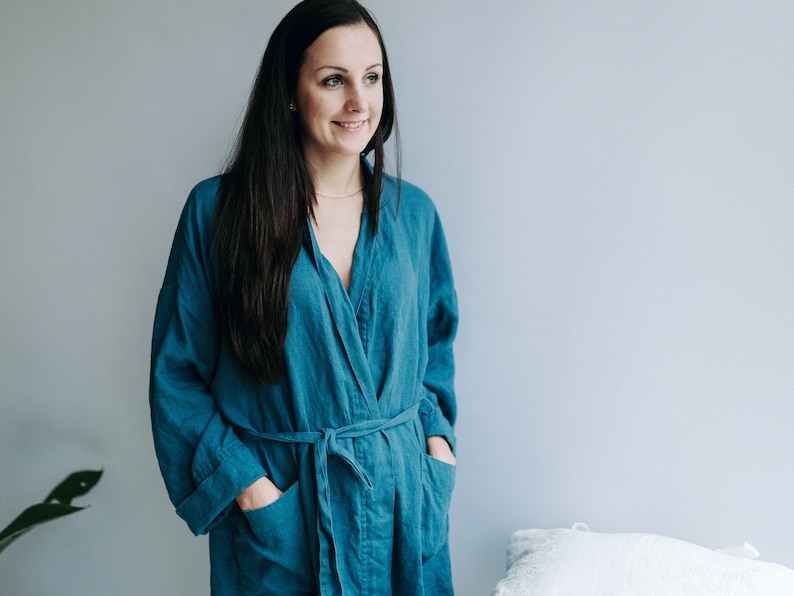May include: A woman wearing a teal blue linen robe with a sash tie. The robe has pockets on the sides.