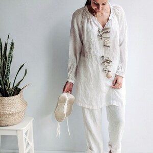 May include: A woman wearing a white linen tunic top and pants with a long, beaded necklace made of driftwood and shells. She is holding a pair of white canvas shoes in her hand.