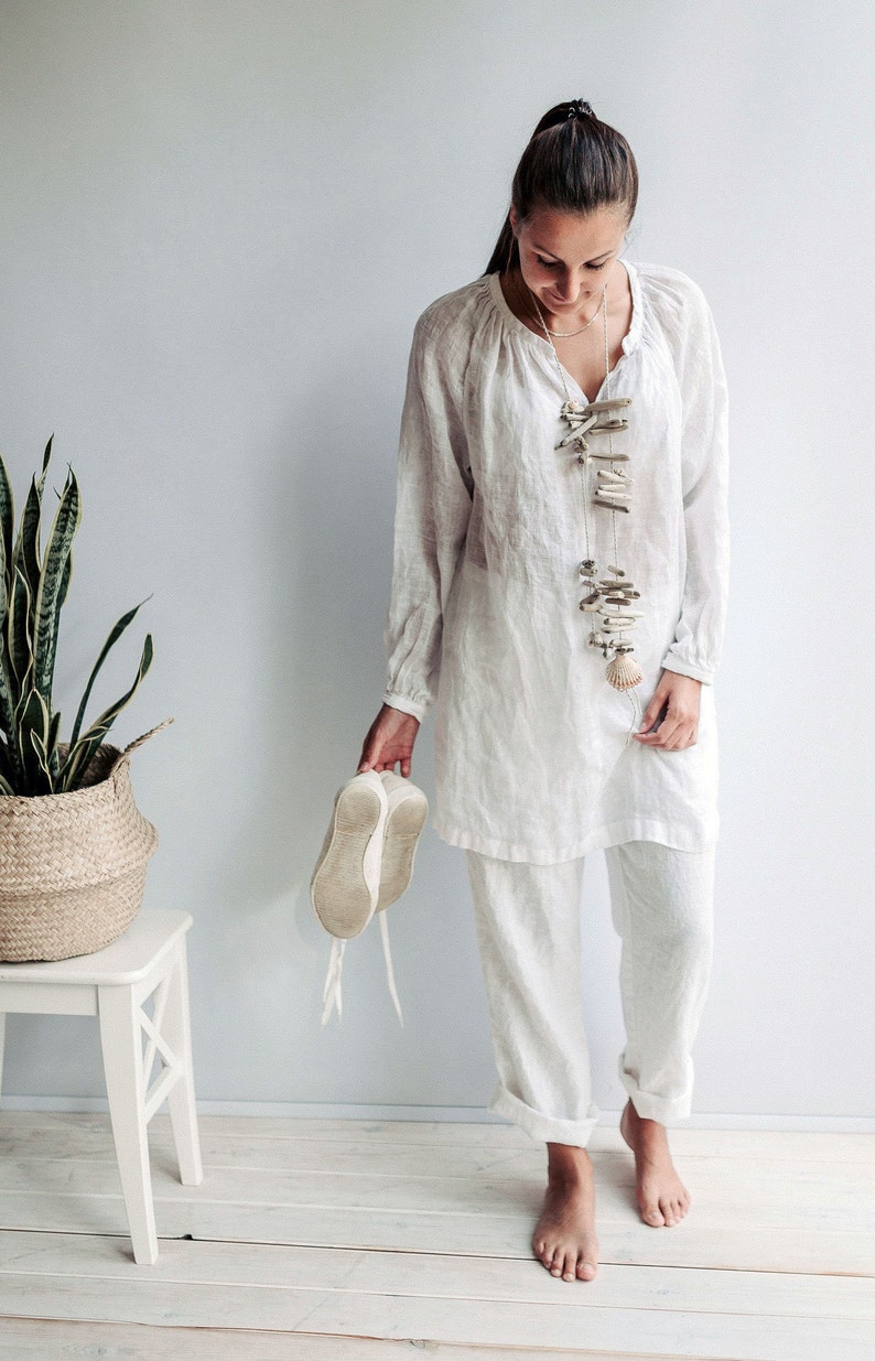 May include: A woman wearing a white linen tunic top and pants with a long, beaded necklace made of driftwood and shells. She is holding a pair of white canvas shoes in her hand.