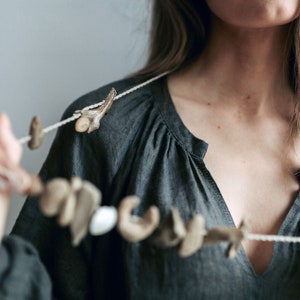 May include: A handmade necklace featuring a string of natural, organic elements in shades of beige and brown. The necklace is composed of various shapes and textures, including what appear to be small shells and bone fragments. The necklace is displayed against a dark gray top.