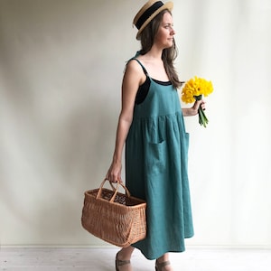 May include: A woman wearing a teal linen dress, a straw hat with a black band, and brown shoes holds a wicker basket and a bouquet of yellow daffodils.