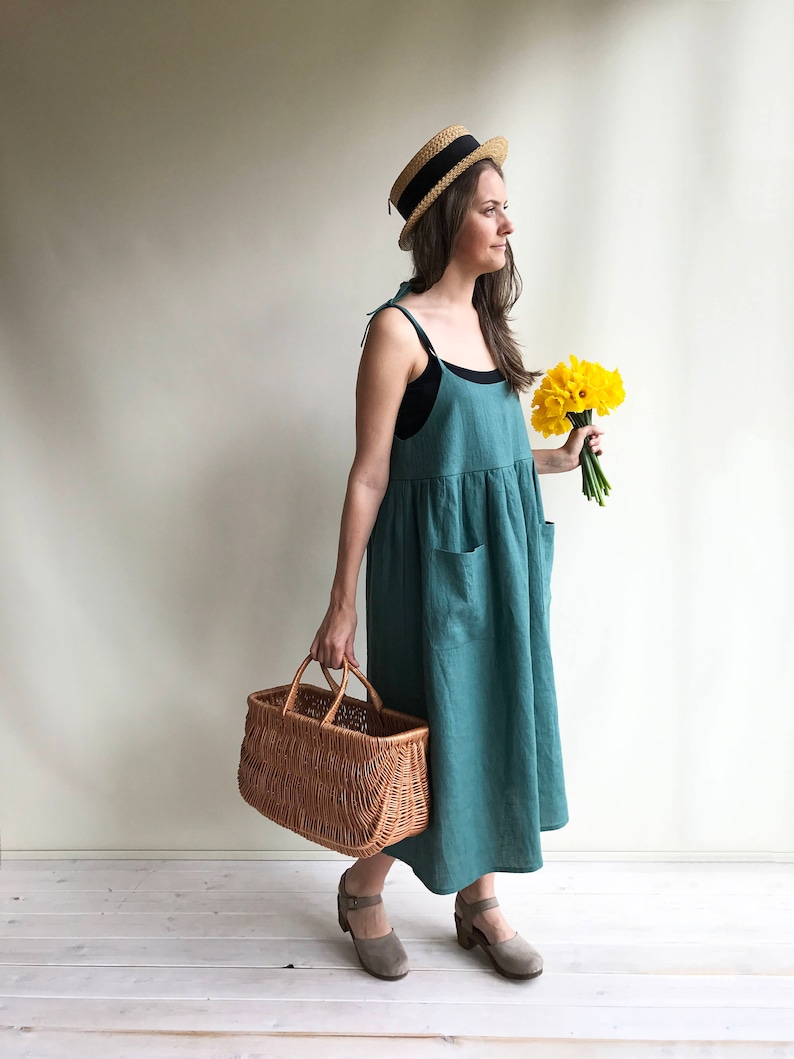 May include: A woman wearing a teal linen dress, a straw hat with a black band, and brown shoes holds a wicker basket and a bouquet of yellow daffodils.
