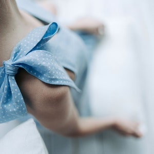 May include: Close-up of a light blue dress with white star-shaped patterns. The dress features a bow detail on the shoulder. The image is focused on the shoulder and arm of the dress.