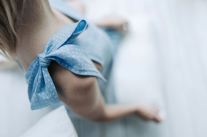 May include: Close-up of a light blue dress with white star-shaped patterns. The dress features a bow detail on the shoulder. The image is focused on the shoulder and arm of the dress.
