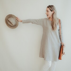 May include: A woman wearing a light gray linen dress with a brown leather shoulder bag and white pants. She is holding a brown hat in her right hand and is looking to the right. She is standing on a brown woven rug.