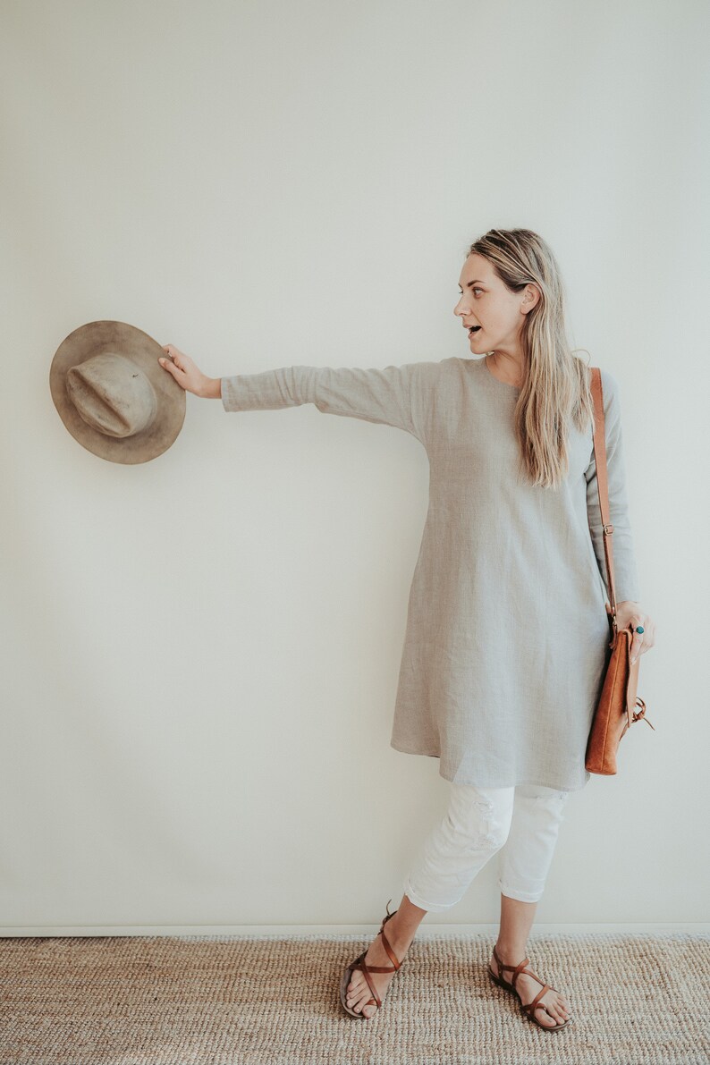 May include: A woman wearing a light gray linen dress with a brown leather shoulder bag and white pants. She is holding a brown hat in her right hand and is looking to the right. She is standing on a brown woven rug.