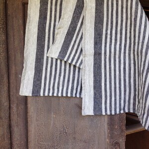 May include: A linen textile with a vertical striped pattern in alternating shades of gray and off-white. The fabric is draped over a wooden surface, showcasing its texture and design. The textile appears to be a towel or a similar home decor item.