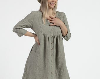May include: A green and white striped linen dress with a button-up front and 3/4 sleeves. The dress has a gathered waist and a relaxed fit. The model is wearing a straw hat.