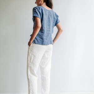 May include: A woman wearing a light blue linen top and white linen pants. She is standing with her hands in her pockets and looking over her shoulder.