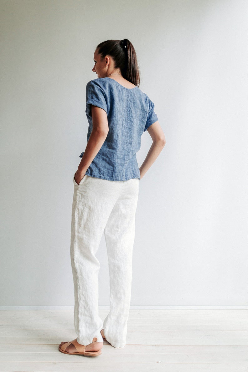 May include: A woman wearing a light blue linen top and white linen pants. She is standing with her hands in her pockets and looking over her shoulder.
