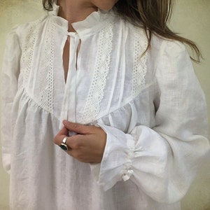 May include: A white linen blouse with a high ruffled collar and long sleeves. The blouse features delicate lace detailing across the yoke and down the front. The sleeves gather at the wrist, finished with small buttons.